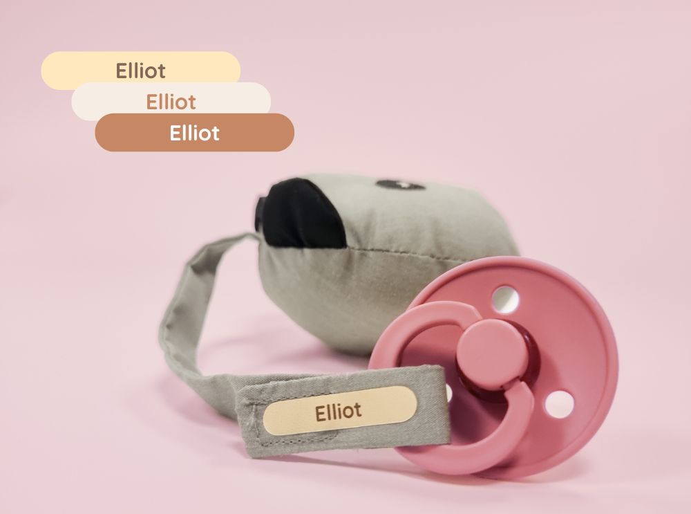 Rosa napp med en personlig namnetikett med texten 'elliot' på ett tygband.