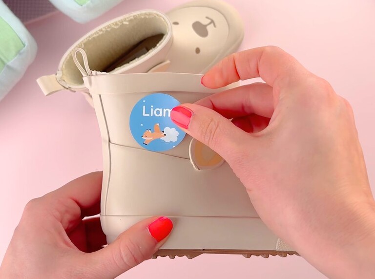 Rund blå namnlapp med rävmotiv fästs på en beige barnstövel