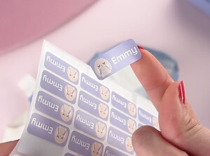 Hand som håller en lila personlig namnlapp med kanindesign och namnet Emmy, dragen från ett klistermärkesark