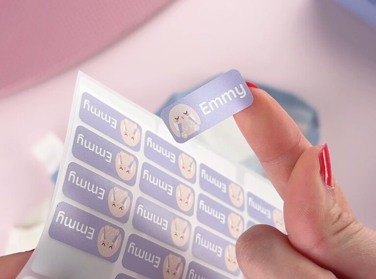 Hand som håller en lila personlig namnlapp med kanindesign och namnet Emmy, dragen från ett klistermärkesark