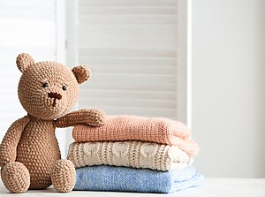 Stickad nallebjörn som sitter bredvid en hög med vikta babykläder i mjuka pastellfärger