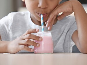 Barn som dricker en rosa smoothie ur ett glas med randigt sugrör vid ett bord