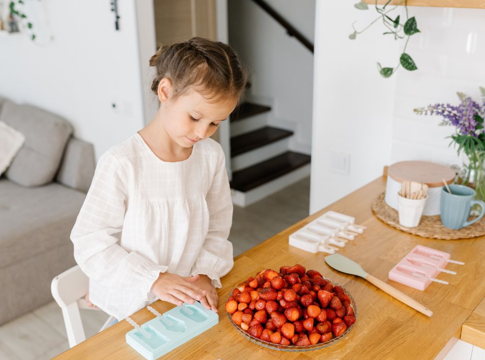 Flicka förbereder jordgubbar vid ett köksbord med glassformar i närheten – redo att göra egna frysta godsaker.
