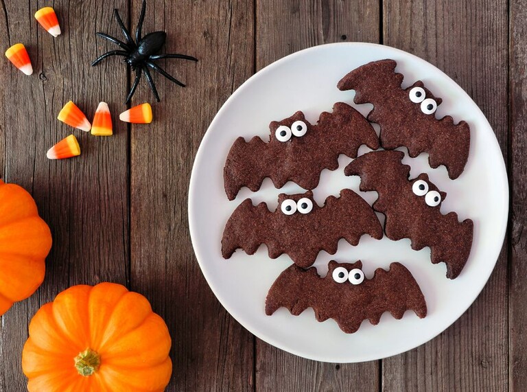 Tallrik med roliga fladdermusformade Halloween-kakor med godisögon, omgivna av pumpor och kandismajs — en perfekt barnvänlig Halloween-godbit