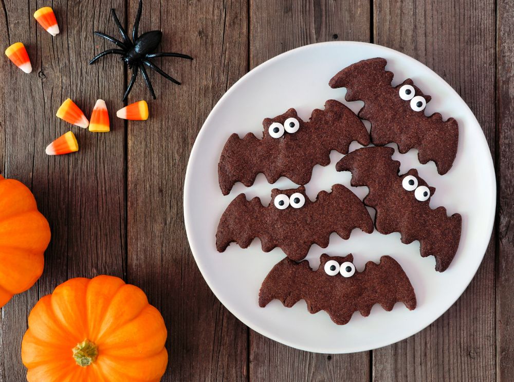 Tallrik med roliga fladdermusformade Halloween-kakor med godisögon, omgivna av pumpor och kandismajs — en perfekt barnvänlig Halloween-godbit