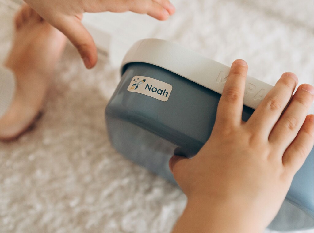 Så förenklar namnlappar vardagen i förskolan 2 Barnhänder håller i en blå matlåda med en namnlapp med texten ”noah”.