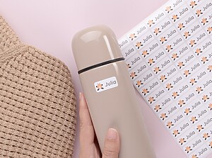 Hand som håller en beige vattenflaska märkt med ett vitt namnklistermärke med lägereldsmotiv och namnet Julia, bredvid ett matchande klistermärkesark