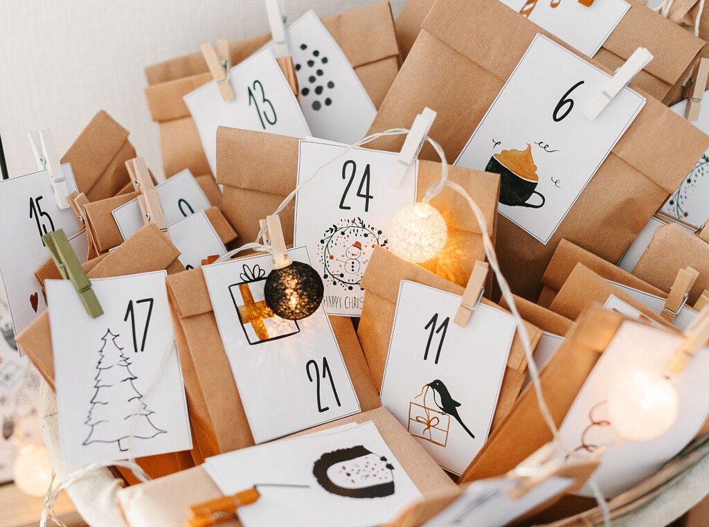 Närbild av en hemmagjord adventskalender med bruna papperspåsar, vita sifferetiketter, klädnypor och en dekorativ ljusslinga.
