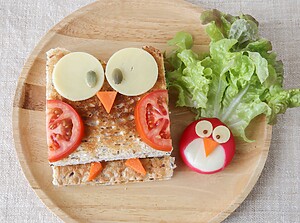 Uggleformad barnsnack av rostat bröd, ost, tomat och morot med ett litet äggkyckling på en träbricka.