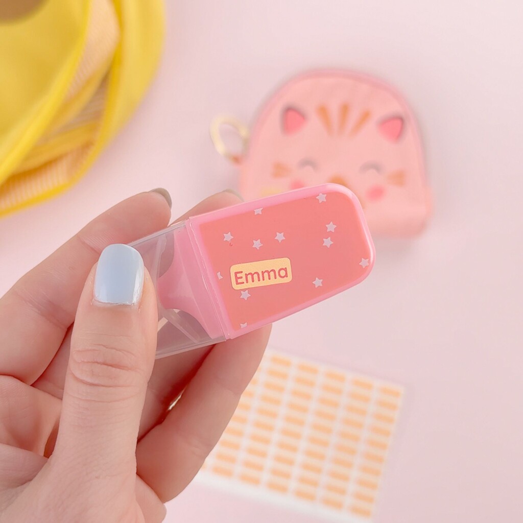 Hand som håller en rosa överstrykningspenna märkt med namnet Emma, med mini-namnlappar i bakgrunden