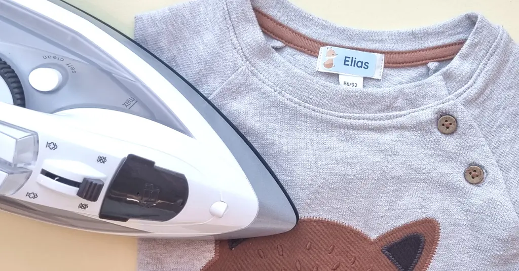 Stryklapp med ett rävmotiv fäst på en grå babytröja, visad bredvid ett strykjärn