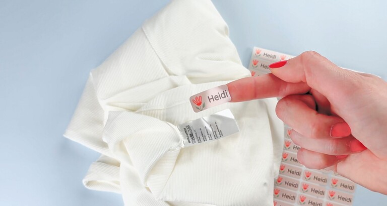 Hand fäster Heidi-namnlapp på tvättrådslapp i vit textil bredvid ark med namnlappar