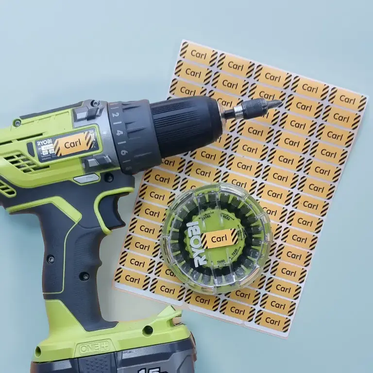 Grön Ryobi-borrmaskin och bitsbox märkta med gula Carl-namnlappar bredvid ark med fler verktygslappar