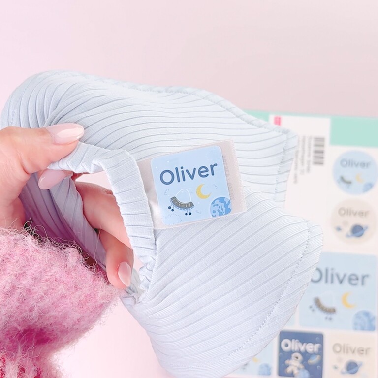 Ljusblå barnmössa märkt med Oliver-namnlapp och ark med stora namnlappar i rymdtema