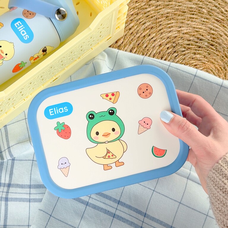 Blå matlåda med ducky-i-grodkostym-design och namnet Elias bredvid dricksflaska i picknickkorg