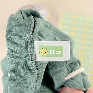 Namnlapp med ducky-motiv fäst på grön barntröja bredvid ark med fler namnlappar