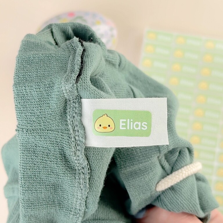 Namnlapp med ducky-motiv fäst på grön barntröja bredvid ark med fler namnlappar