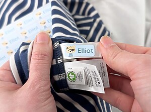 Hand applying a blue name label with the name Elliot to a clothing tag inside a striped shirt