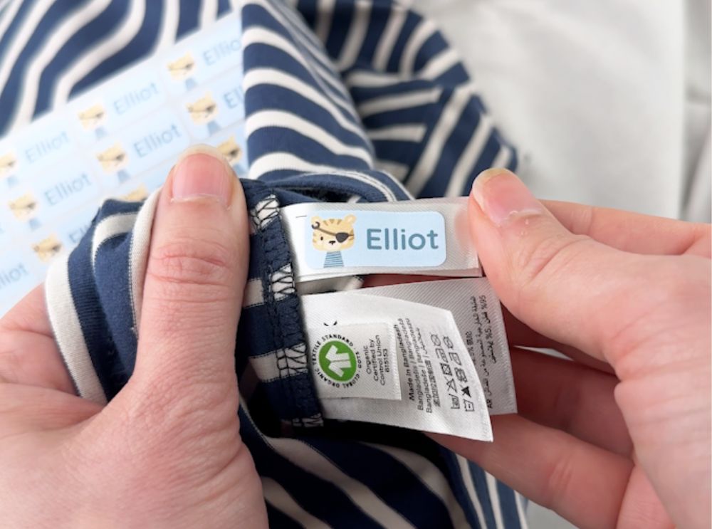 Hand applying a blue name label with the name Elliot to a clothing tag inside a striped shirt