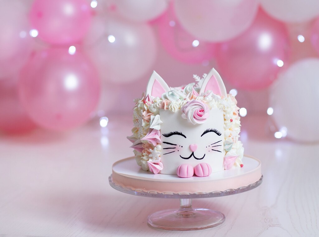Kattformad födelsedagstårta på tårtfat med rosa ballonger i bakgrunden
